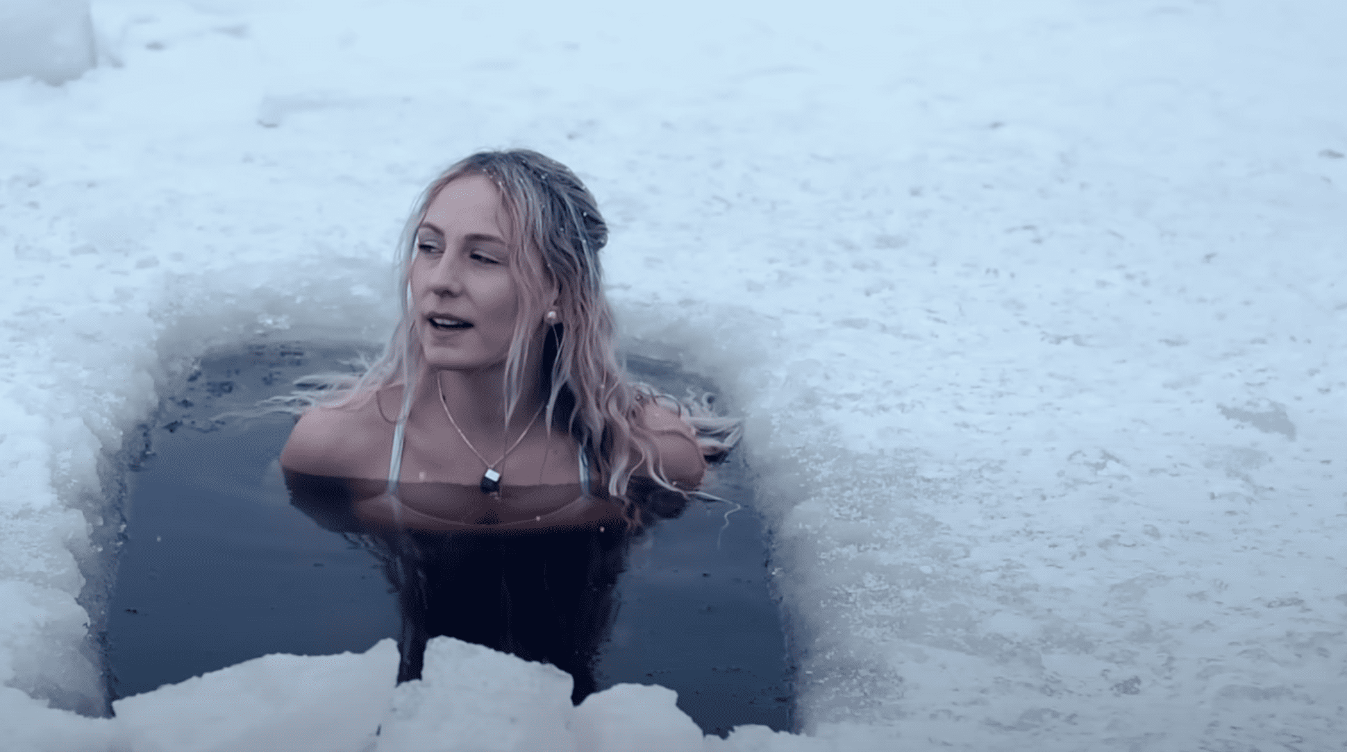 a hole in the ice of a lake. in it sits a woman, only her shoulders and head are above the water.